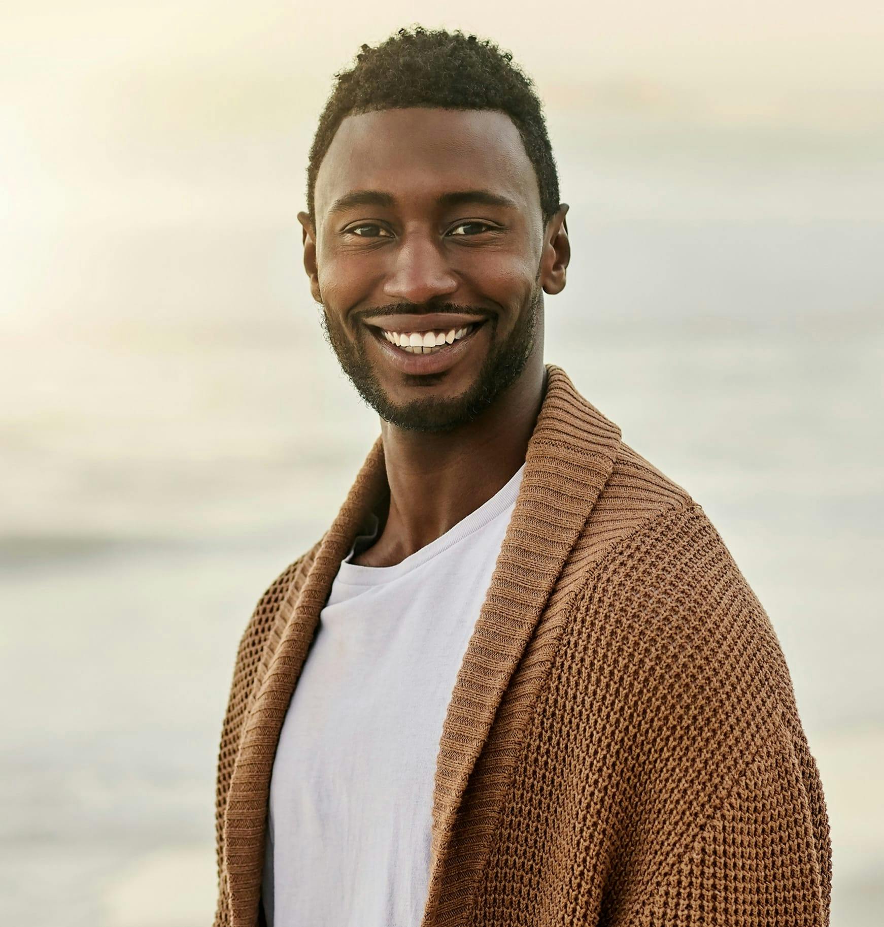 smiling man in brown cardigan standing on beach with ocean in background