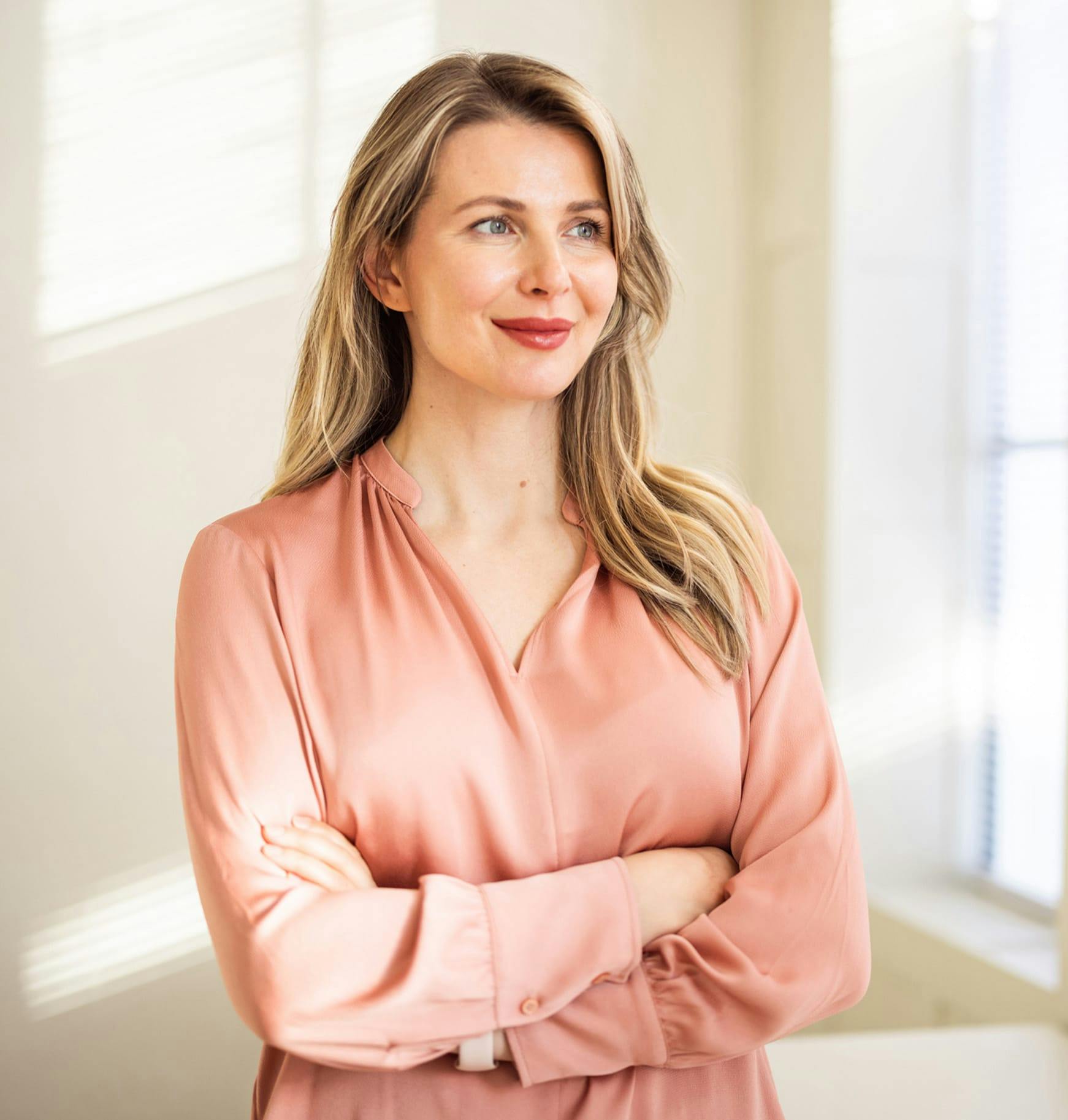 smiling blond woman in pink shirt standing in front of window with arms crossed