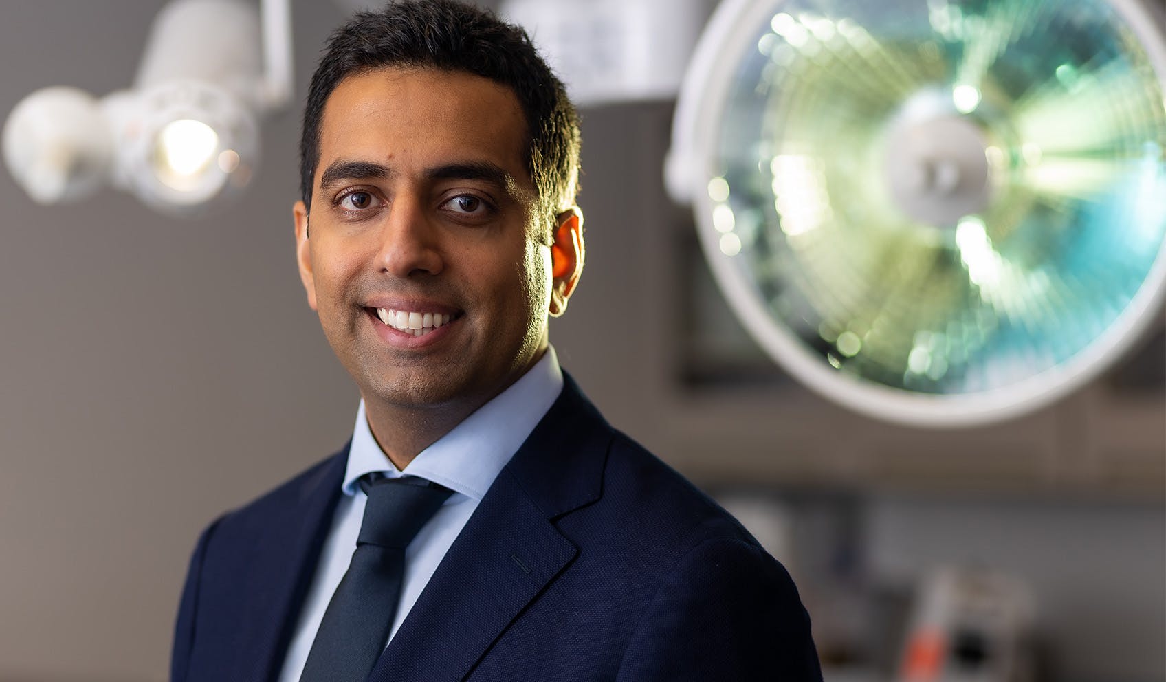 Dr. Akshay Sanan Smiling in suit standing in surgery room