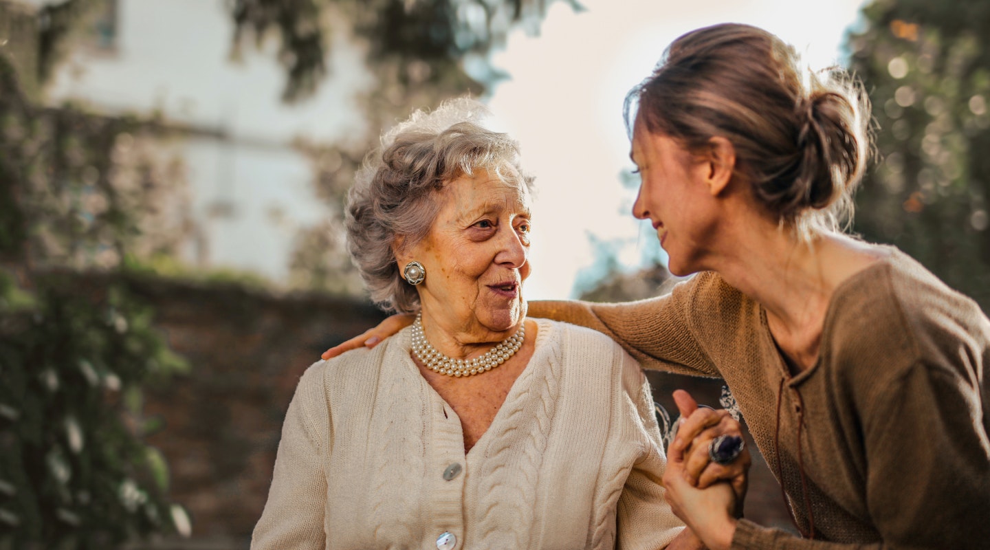 Arc Health - GP care for rest homes and aged residential care. A younger women talking with an older women with her arm around her. They are in a garden setting with nice light. They are both smiling at each other.