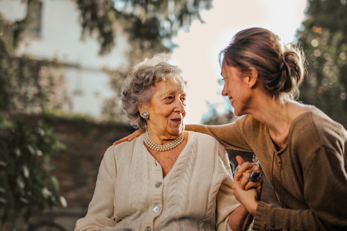 A younger women talking with an older women with her arm around her. They are in a garden setting with nice light. They are both smiling at each other.