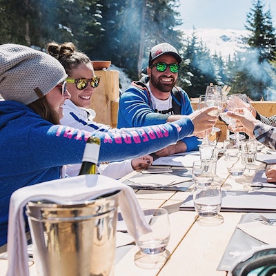 A group of people cheerfully toasts around a large outdoor table in the mountains under the sun.