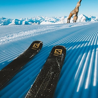 Deux skis noirs reposent sur une piste fraîchement damée, en montagne. Au loin, une grande statue de bouquetin se dresse devant un panorama de sommets enneigés sous un ciel bleu clair.