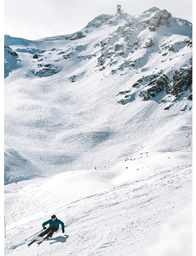 Un skieur descend une large piste enneigée au pied d’une montagne rocheuse, sous un ciel légèrement nuageux.