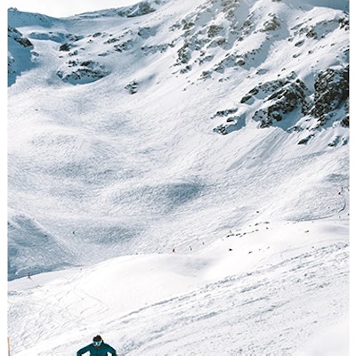 Un skieur descend une large piste enneigée au pied d’une montagne rocheuse, sous un ciel légèrement nuageux.