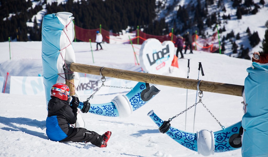 un enfant portant un casque rouge, soufflant dans une corne, est assis sur la neige à côté de deux balançoires décorées, dans un environnement de station de ski. Les montagnes sont visibles en arrière-plan.