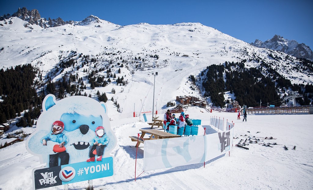 Zone de repos en plein air sur une piste de ski, une grande mascotte bleue en forme de Yooni, notre yéti préféré, blanc avec des petites cornes bleues. Montagnes enneigées en arrière-plan.
