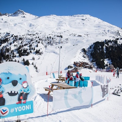 Outdoor rest area on a ski slope, featuring a large blue mascot shaped like Yooni, our favourite yeti, white with small blue horns. Snow-capped mountains in the background.
