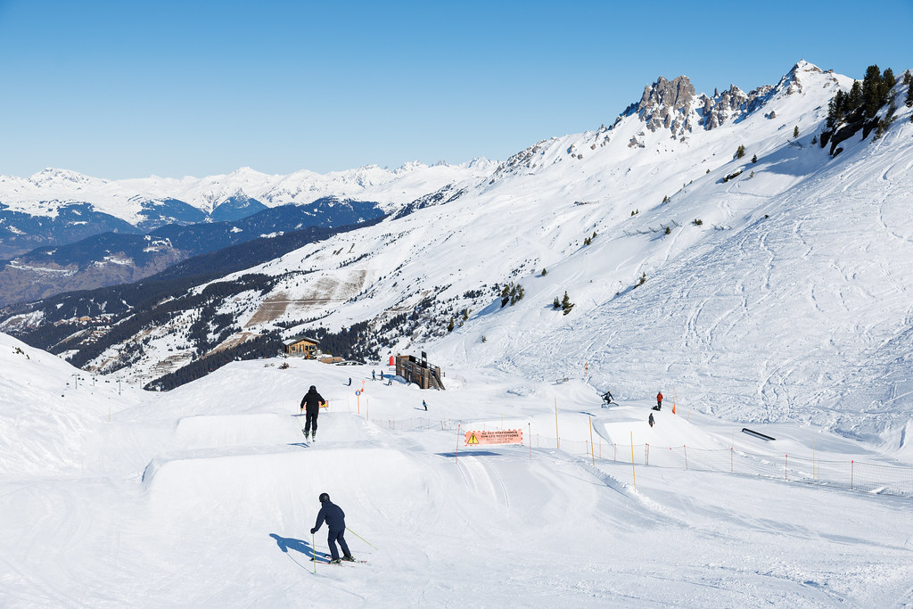 Deux skieurs descendent une ligne de sauts dans un snowpark, entourés de montagnes enneigées sous un ciel bleu.