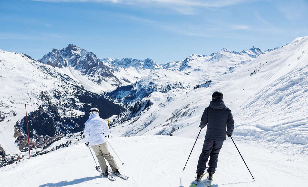 2 skieurs dévalent une piste qui surplombe la station de Mottaret, avec ses batiments en bois et face à une chaine de montagne enneigée, sous un ciel bleu