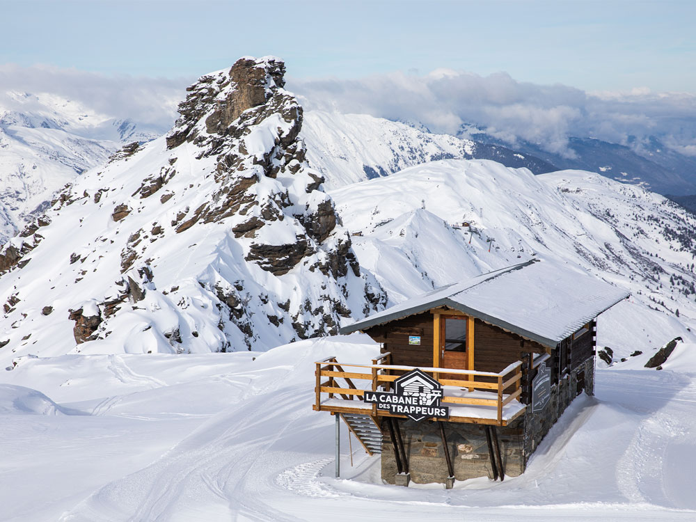 Un chalet en bois dans un paysage de montagne enneigé. À l’arrière, une montagne rocheuse. Le ciel est clair, . Sur le balcon du chalet, une pancarte indique : "La cabane des trappeurs".