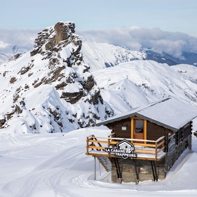 A wooden chalet in a snow-covered mountain landscape. Behind it, a rocky mountain. The sky is clear. On the balcony of the chalet, a sign reads: ‘La cabane des trappeurs".