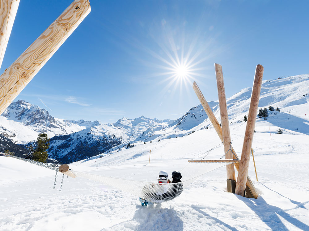 dans un payasge enneigé et sous un ciel bleu ensoleillé, 2 personnes sont installées dans unhamac géant, face aux montagnes