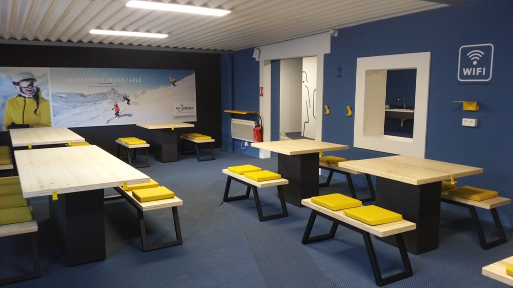Salle de pause moderne avec tables et bancs en bois, coussins jaunes. Murs bleus, grande photo de skieurs, accès Wi-Fi indiqué.