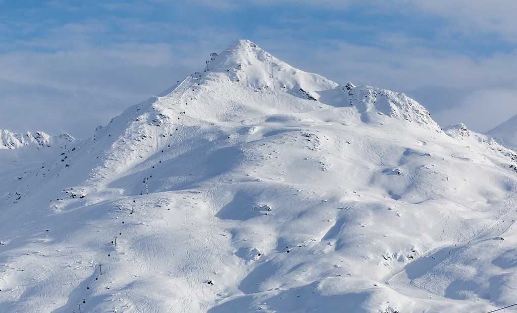 Montagne enneigée avec remontée mécanique et traces de ski sur les pentes. Ciel bleu partiellement nuageux