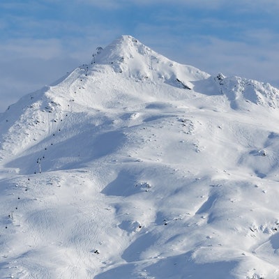 Snow-covered mountain with ski lift and ski tracks on the slopes. Partly cloudy blue sky.