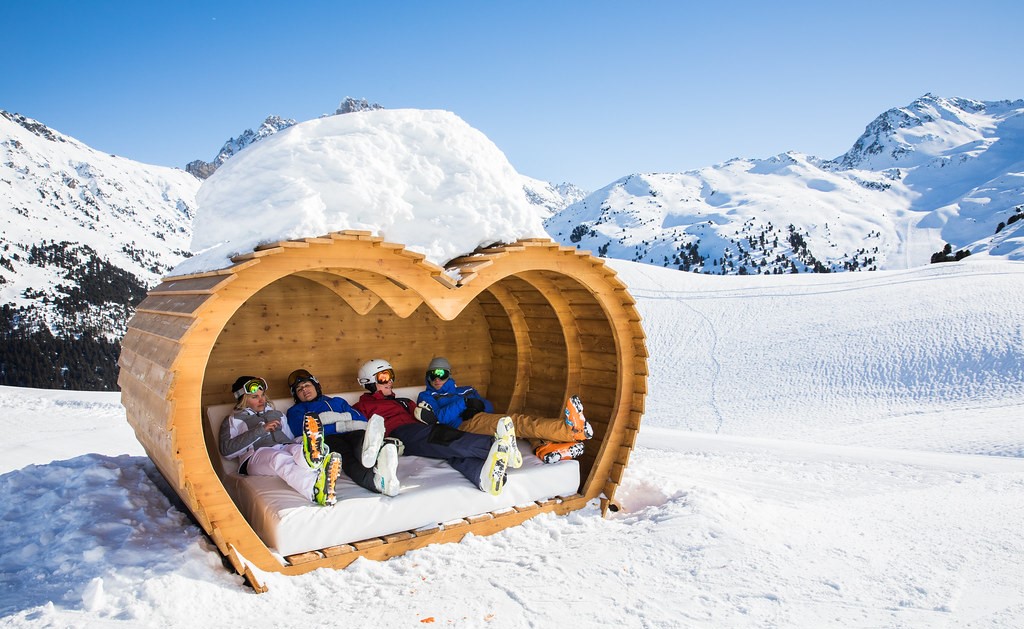 Structure en bois en forme de coeur, à l'intérieur 4 skieurs se détentes sur une banquette blanche, dans un décor ennéigé et sous un ciel ensoleillé