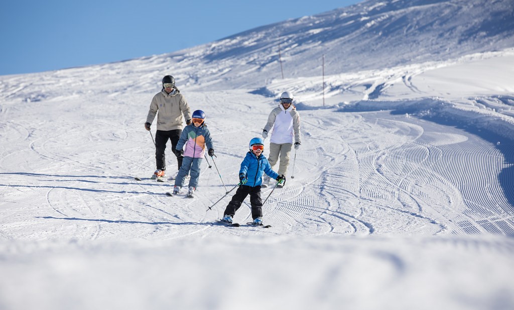 une famille de 4 personnes derscendent une piste de ski, en premier les enfants et derrière les adultes, sous un ciel dégagé