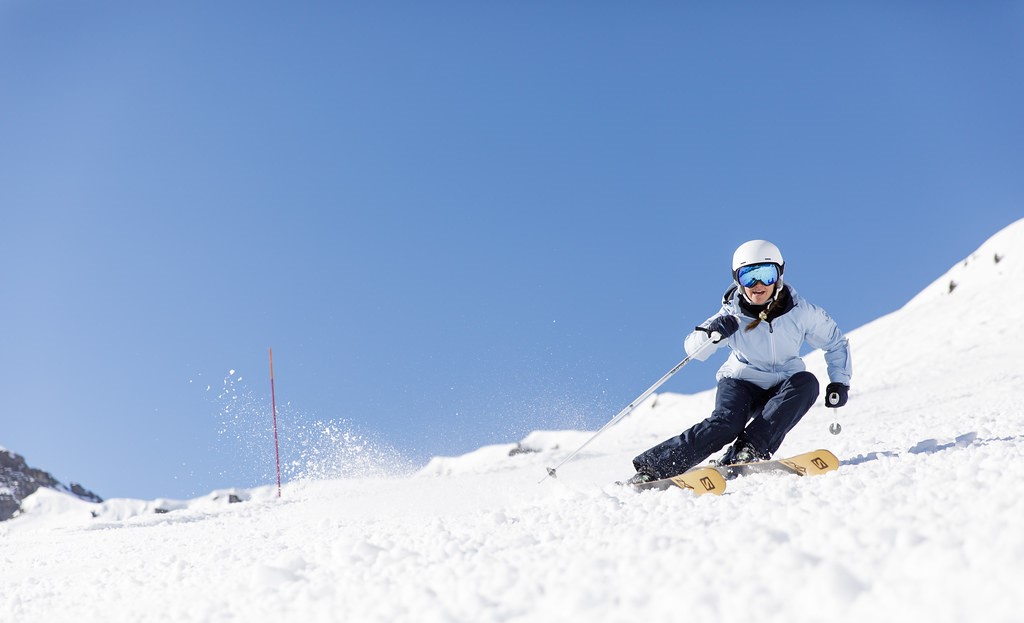 A skier wearing a white helmet, sky blue ski jacket and navy blue trousers skiing down a slope with a smile, under clear sky