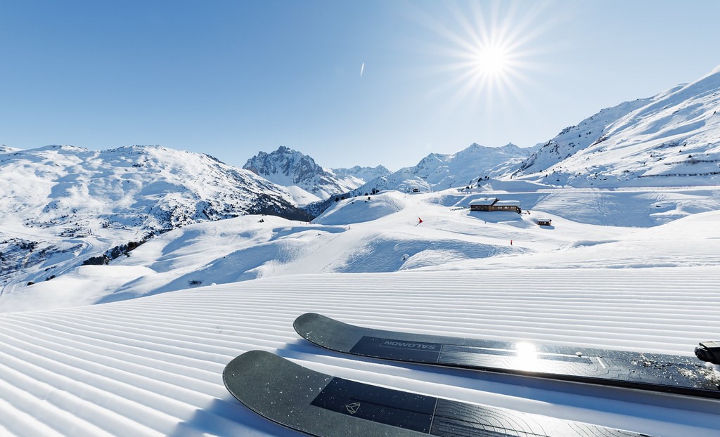 Deux skis posés sur une piste damée sous un grand soleil. Au loin, des montagnes enneigées, un ciel bleu clair et un chalet en bois sur la droite.