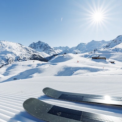 Deux skis posés sur une piste damée sous un grand soleil. Au loin, des montagnes enneigées, un ciel bleu clair et un chalet en bois sur la droite.