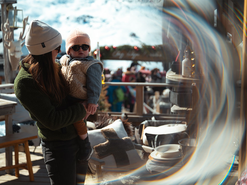 Un jeune enfant avec un bonnet rose claire et des lunettes de soleil, se fait porter par une femme dans un restaurant dans Les 3 Vallées