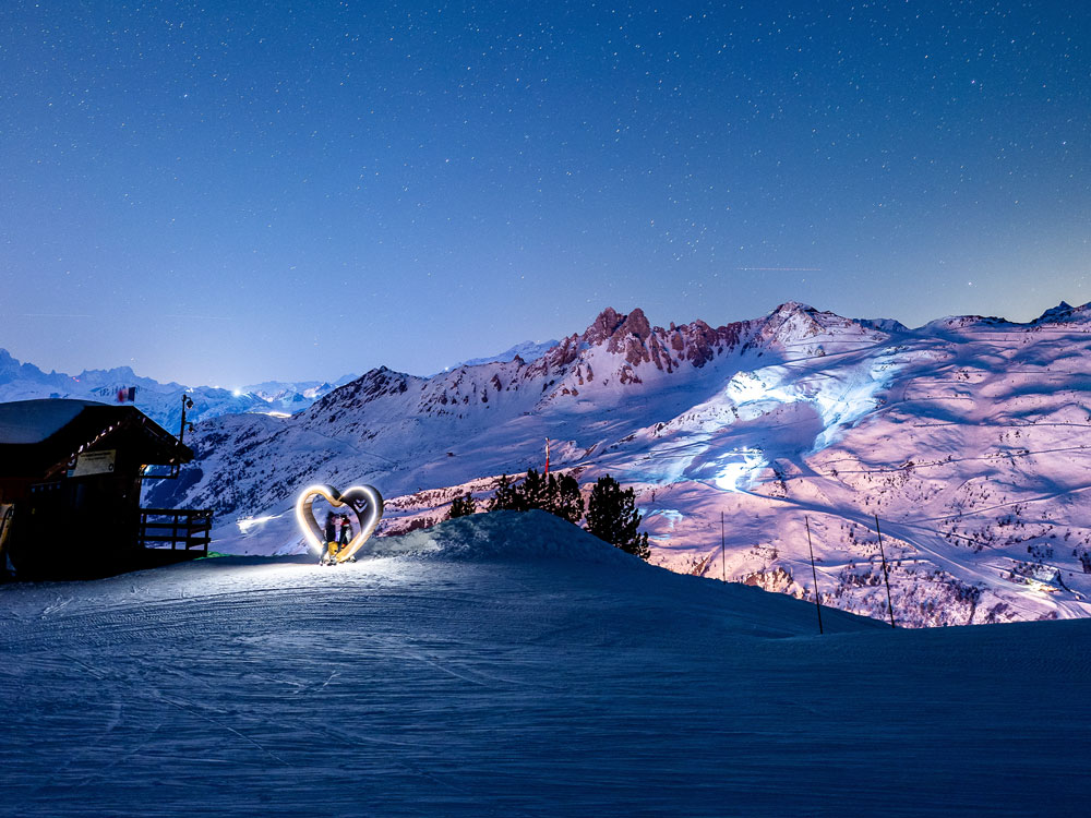 Sous un ciel étoilé d'hiver, un coeur en bois illuminé abrite 2 personnes, derrière montagne enneigée éclairée