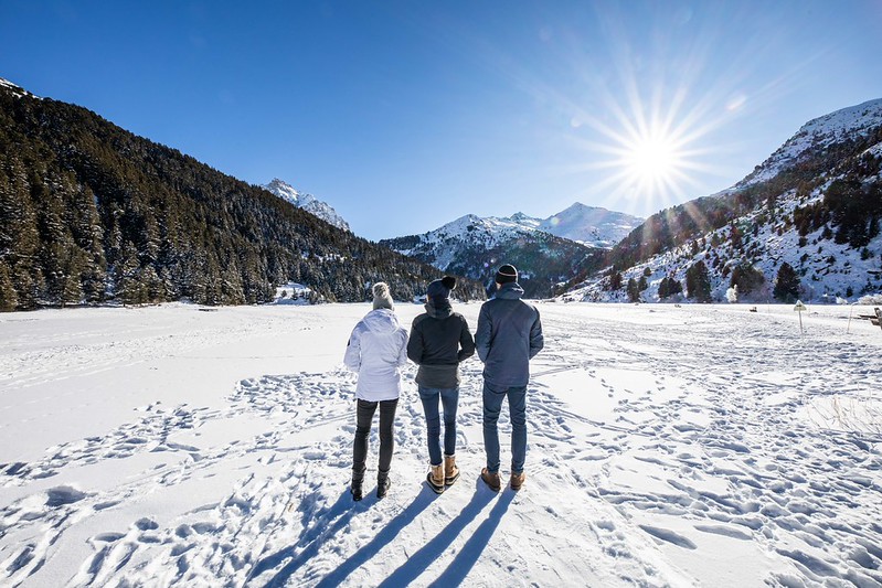 3 personnes de dos devant une étendu de neige et entourés de monatge sous un beau ciel bleu