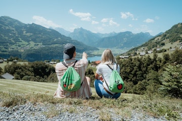 Eine Wanderin und ein Wanderer sitzen auf einer Wiese, fotografiert von hinten mit Blick über ein Tal mit einem See