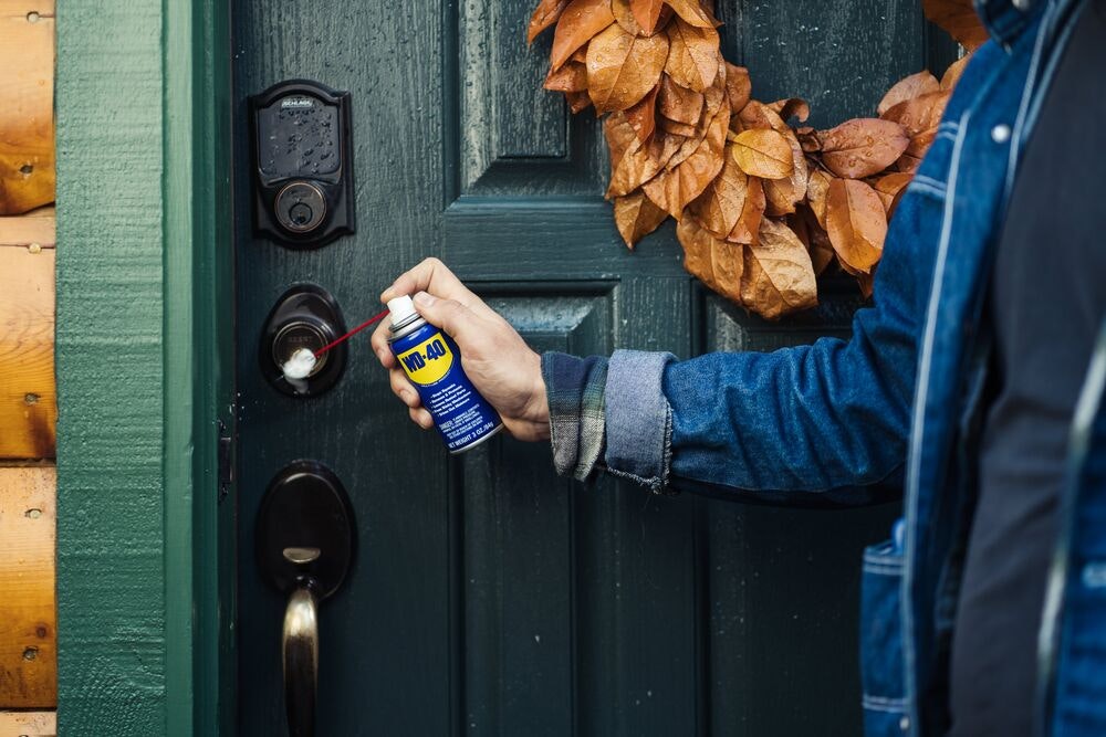 Hand spraying WD-40 into a front door lock on a dark green door with a leaf wreath.