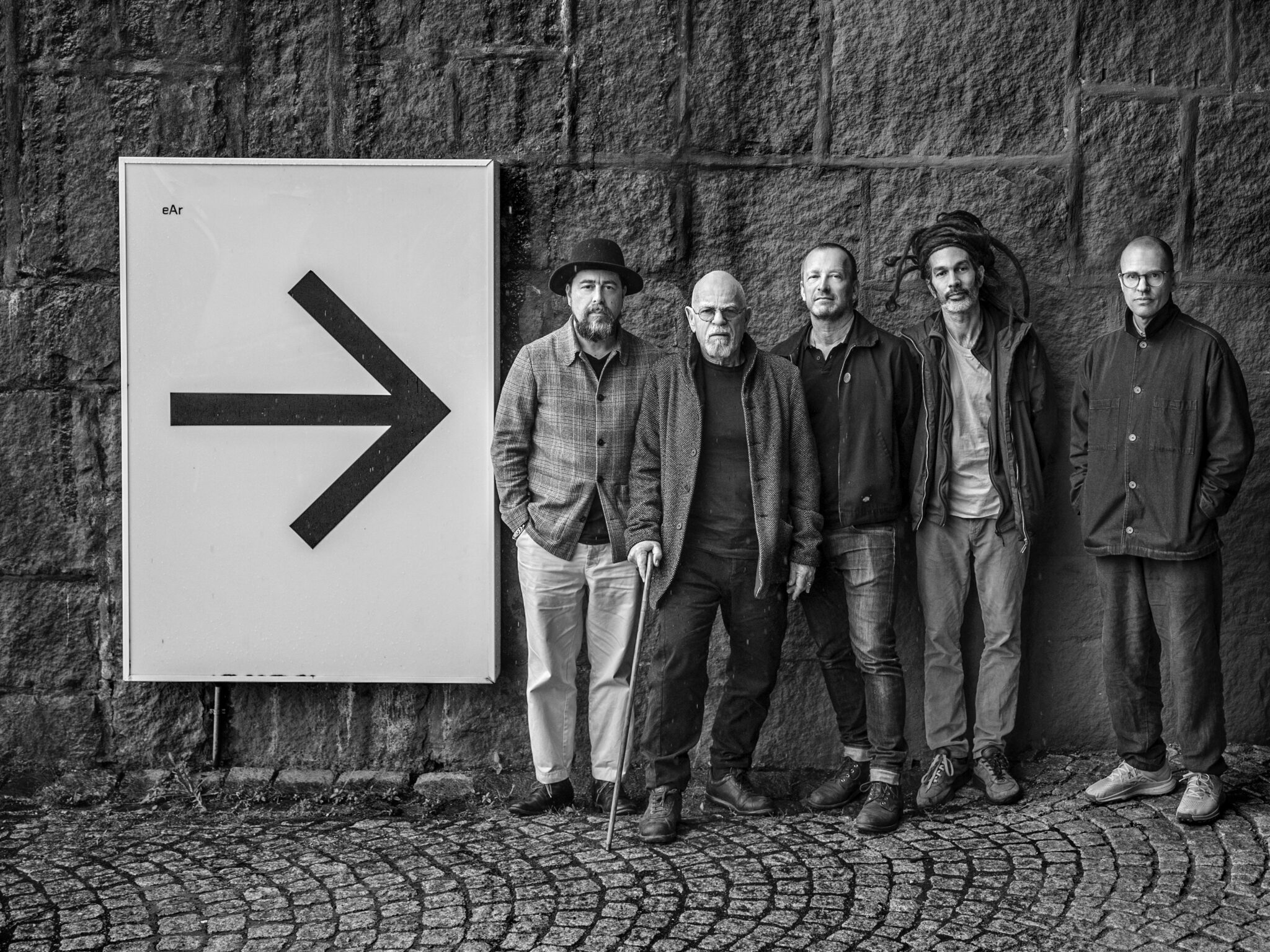 Svartvit bild som visar fem personer som står på kullersten. En stor pil pekar på dem från vänster.