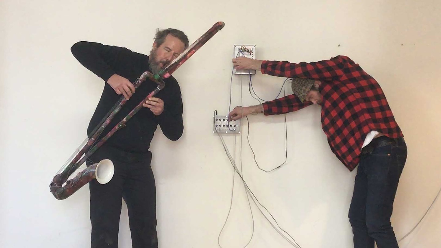 På bilden syns en person med ett blåsinstrument och en person som står böjd med sladdar mot en vägg.