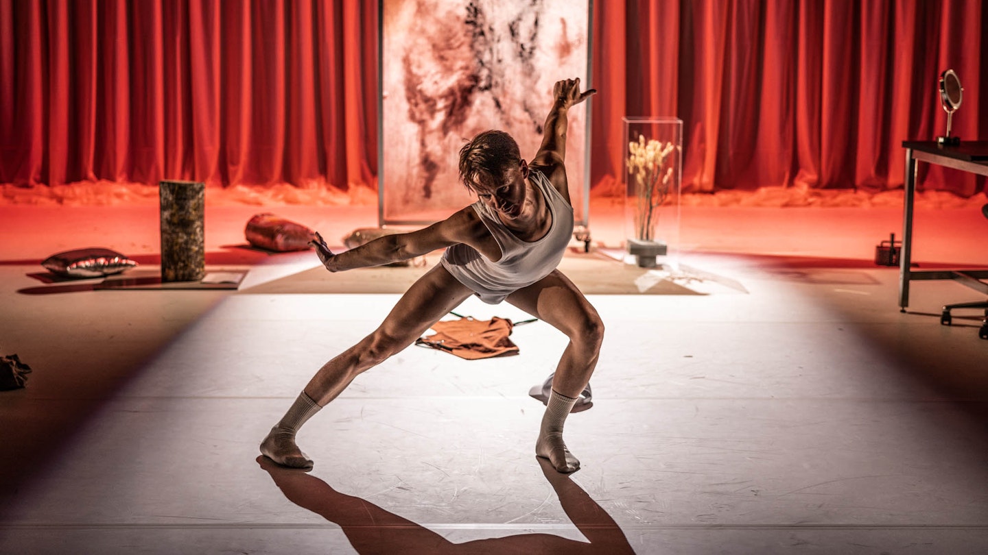 Föreställningsfoto från "Mössan". Foto: Mats Bäcker Skådespelare och dansare Olof Yassin i en rörelse. I bakgrunden syns en skärm med abstrakt motiv, samt röda draperier.
