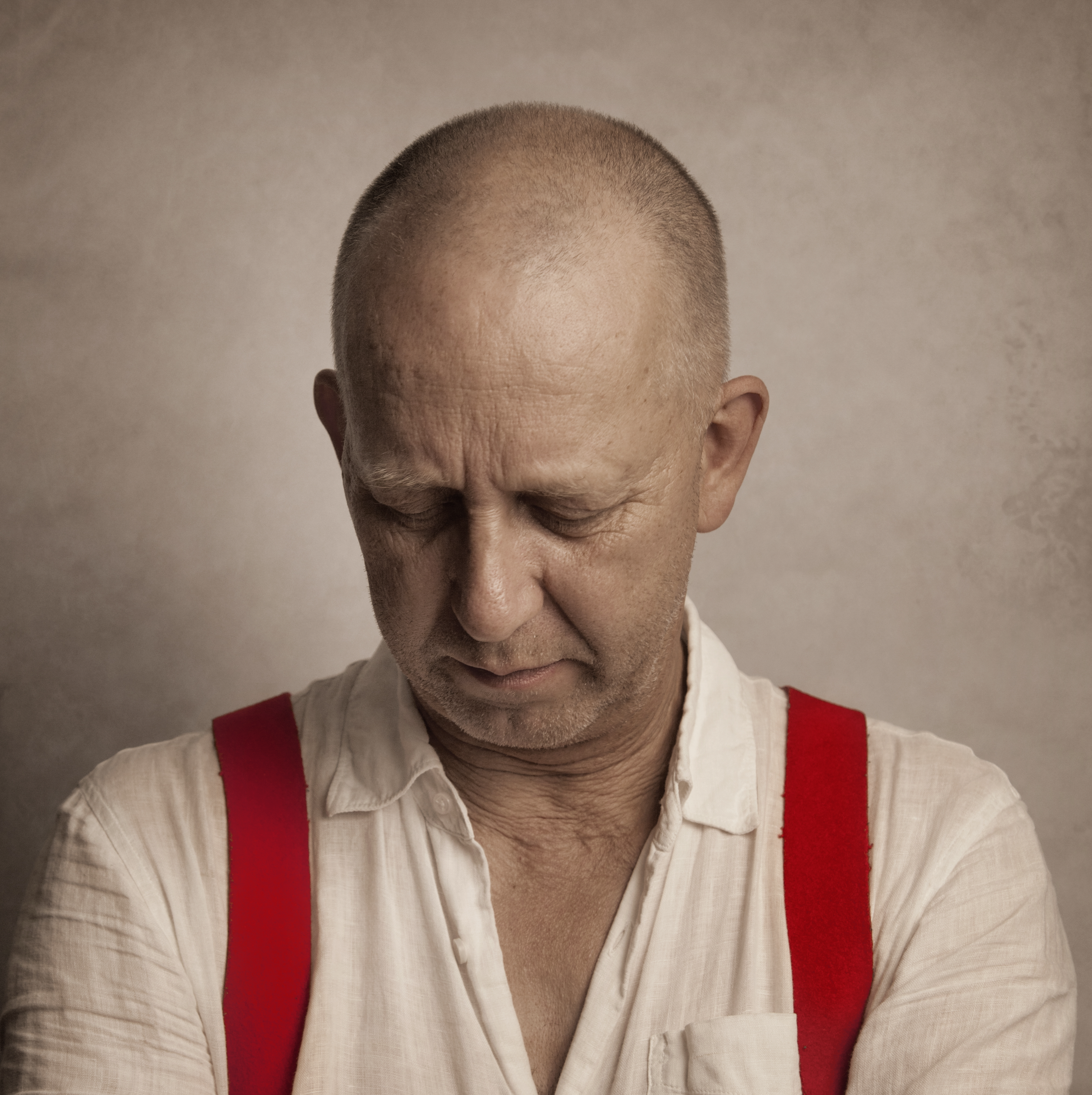 Bilden visar artisten som tittar nedåt. På sig har han en vit skjorta och röda hängslen.