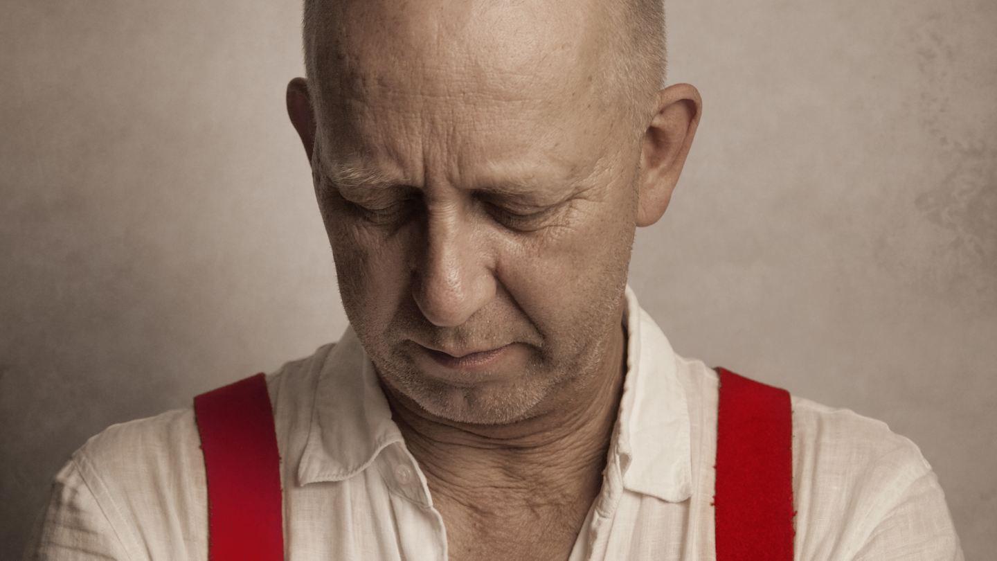 Bilden visar artisten som tittar nedåt. På sig har han en vit skjorta och röda hängslen.