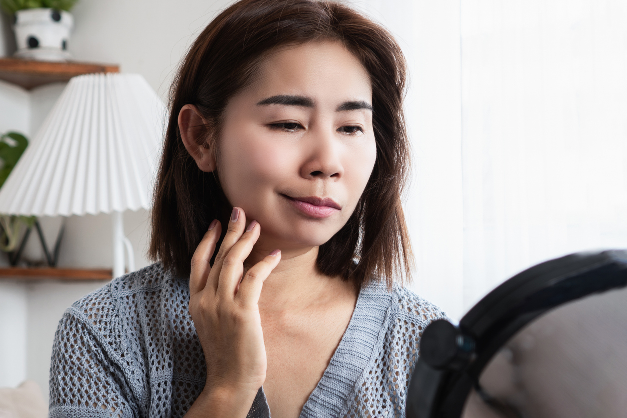 Woman gently touching her cheek while looking into a handheld mirror.