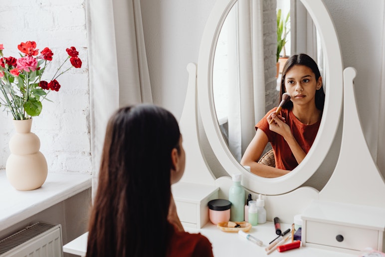 A teenage girl sits at a dressing table applying makeup with a brush while looking at her reflection in an oval mirror.