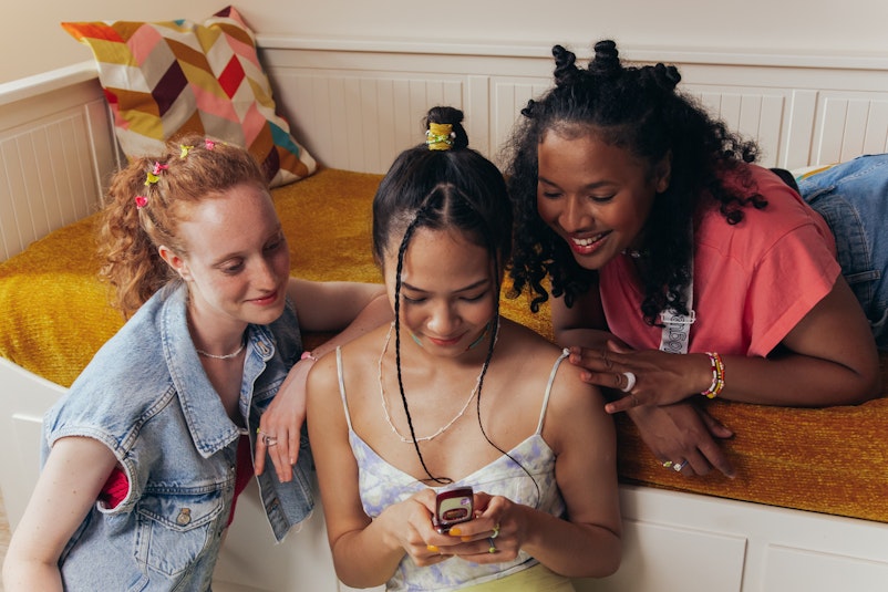 Three teenage girls with braided hair, hair clips, and beaded jewellery lean together looking at a phone one of them is holding.