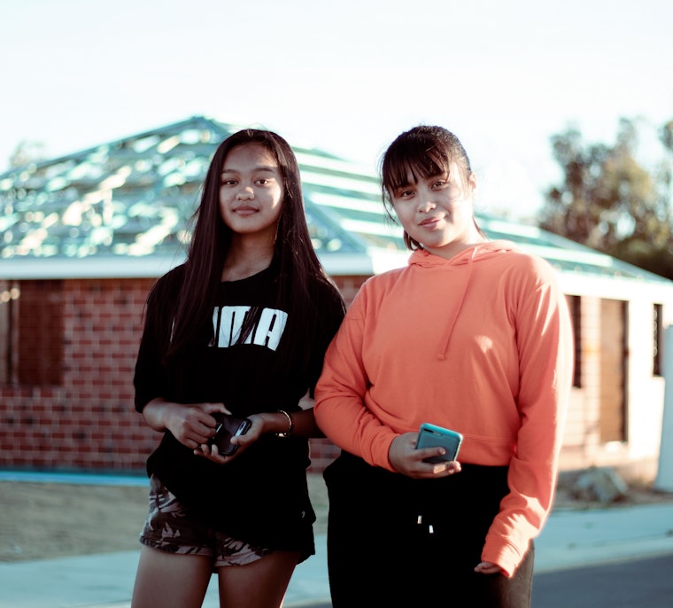 Two teenage girls stand outside near a house under construction, both holding phones and smiling at the camera.