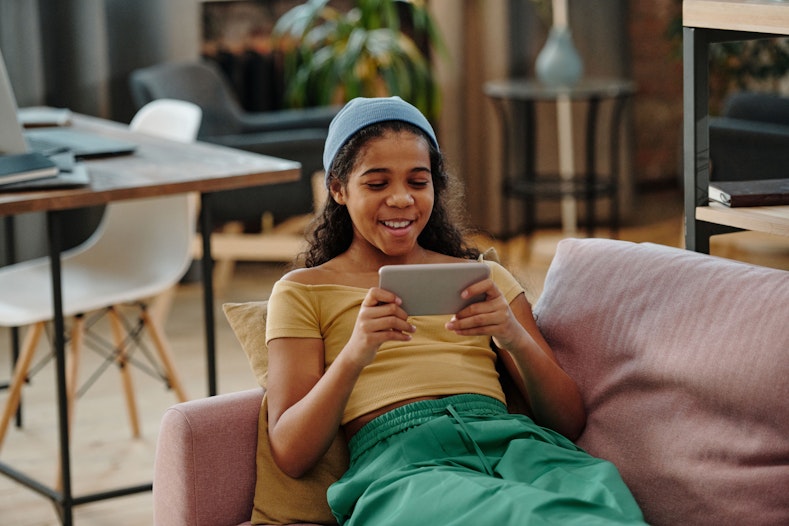 A teenage girl in a beanie sits on a couch smiling while looking at her phone held horizontally in both hands.