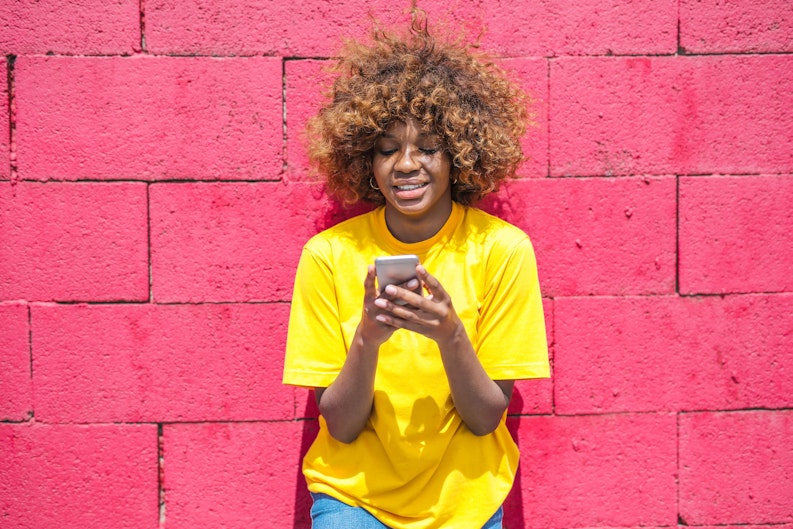 A teenage girl with curly hair stands against a bright pink brick wall, holding a phone.