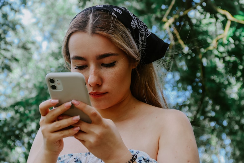 A teenage girl with a headscarf stands outdoors among trees, looking at her phone with a concerned expression.