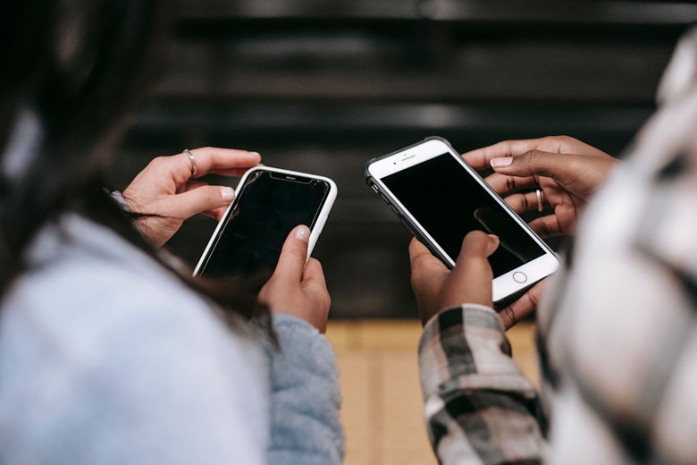 Two people stand close together, each holding a smartphone with a dark screen, seen from behind over their shoulders.