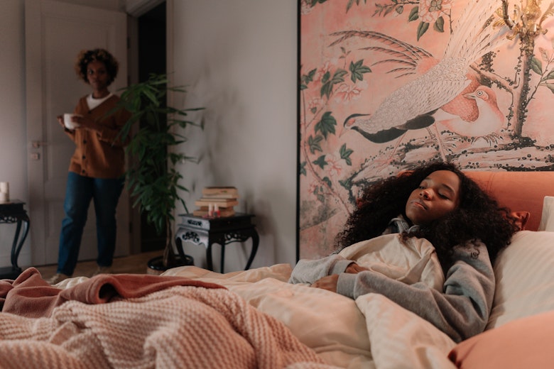 A teenage girl lies in bed under blankets looking tired while a woman stands in the background holding a cup.