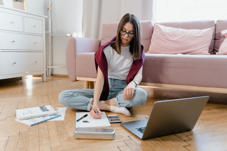 A teenage girl in glasses sits cross-legged on the floor writing on paper, with a laptop, calculator, and textbooks spread around her.