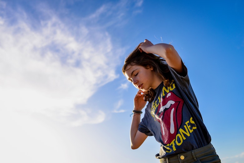 A teenager stands against a bright blue sky with wispy clouds, wearing a Rolling Stones graphic t-shirt and running a hand through their hair.