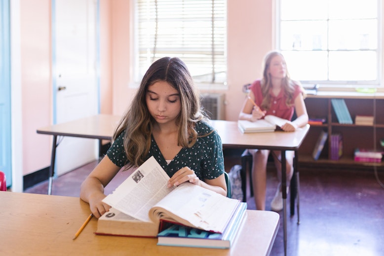 A girl reads from an open textbook at a classroom desk while another girl sits at a desk in the background looking away.