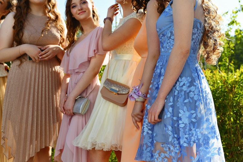 A group of teenage girls in pastel-coloured formal dresses stand together outdoors, smiling.
