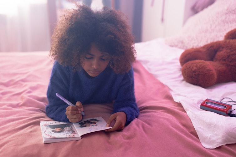 A young girl lies on her bed writing in a diary with a photo tucked inside, with a teddy bear beside her.