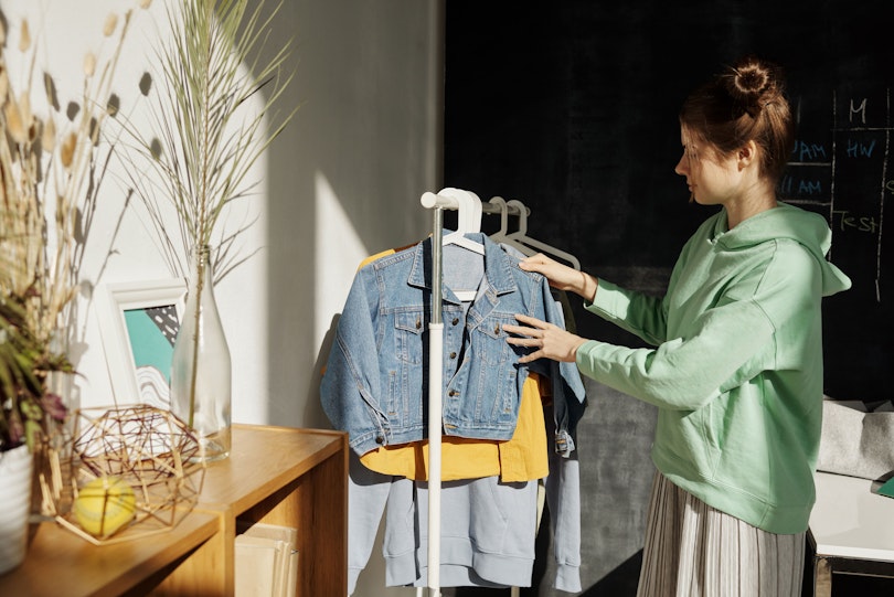 A teenage girl looks through denim jackets and hoodies hanging on a clothing rack in her bedroom.